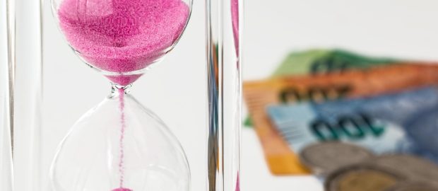 Klepsydra z różowym piaskiem obok banknotów.