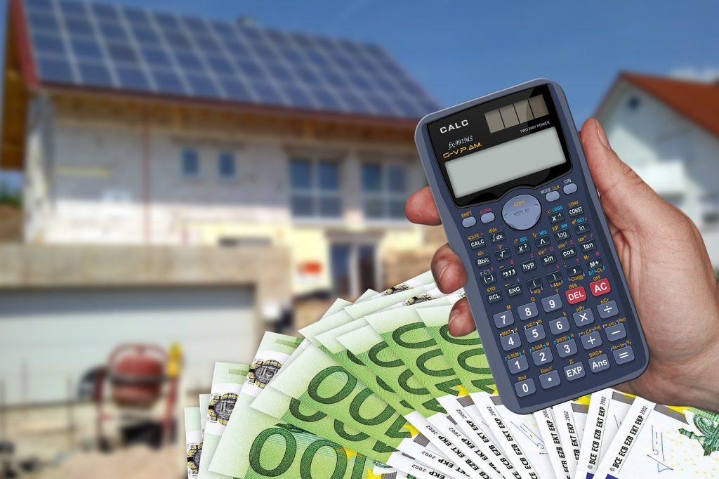 Dłoń trzyma kalkulator i banknoty euro na tle domu z instalacją fotowoltaiczną.