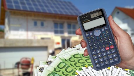 Dłoń trzyma kalkulator i banknoty euro na tle domu z instalacją fotowoltaiczną.