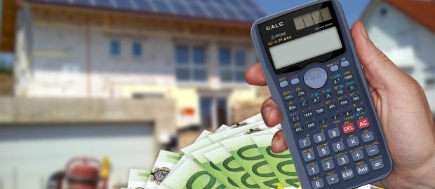 Dłoń trzyma kalkulator i banknoty euro na tle domu z instalacją fotowoltaiczną.