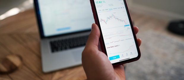 Dłoń trzymająca telefon z wykresem notowań giełdowych na tle laptopa z platformą inwestycyjną.