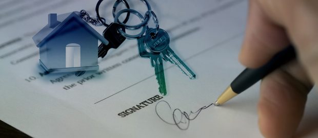 Podpisywanie dokumentu umowy z kluczami i miniaturą domu w tle.